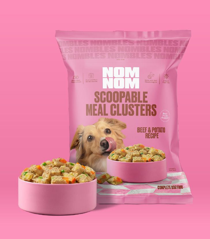 Nom Nom Scoopable Meal Clusters in pink packaging feature the Beef & Potato Recipe,
with a golden-colored dog pictured licking its nose. The bag highlights that the clusters
are complete dog food and can be poured from frozen for easy serving. Two pink bowls in
front show scoopable, bite-sized clusters with visible mix-ins, including orange and green
vegetable pieces, on a clean pink background.